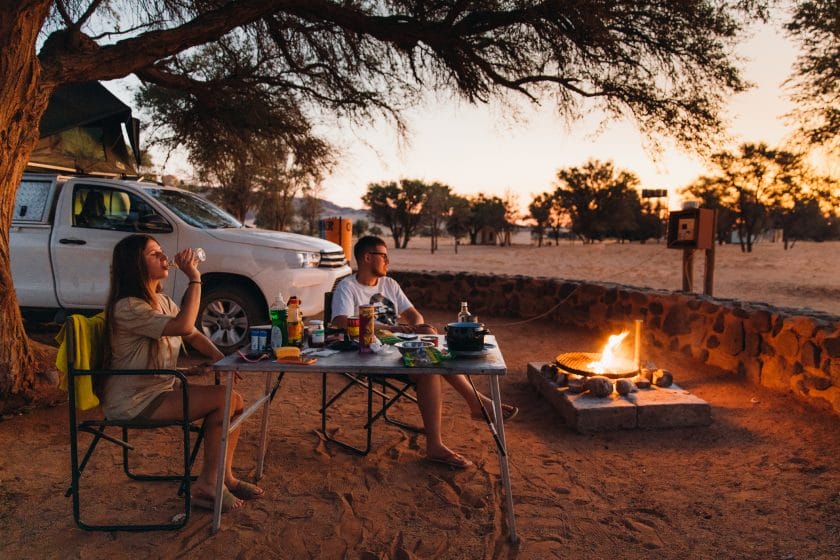 Woman and man traveler camping with a camper car under the tree during sunset in the desert of Namibia